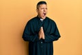 Young latin priest man praying with hands together angry and mad screaming frustrated and furious, shouting with anger