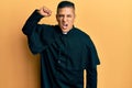 Young latin priest man standing over yellow background angry and mad raising fist frustrated and furious while shouting with anger