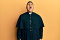 Young latin priest man standing over yellow background angry and mad screaming frustrated and furious, shouting with anger