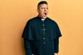 Young latin priest man standing over yellow background angry and mad screaming frustrated and furious, shouting with anger