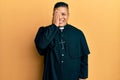 Young latin priest man standing over yellow background covering one eye with hand, confident smile on face and surprise emotion Royalty Free Stock Photo