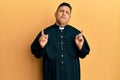 Young latin priest man standing over yellow background pointing up looking sad and upset, indicating direction with fingers,