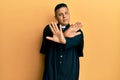Young latin priest man standing over yellow background rejection expression crossing arms and palms doing negative sign, angry