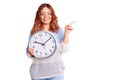 Young latin woman holding big clock smiling happy pointing with hand and finger to the side Royalty Free Stock Photo
