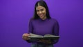 Young latin woman holding book open and reading in purple studio; learning knowledge curiosity focus Royalty Free Stock Photo