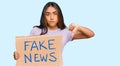 Young latin woman holding fake news banner with angry face, negative sign showing dislike with thumbs down, rejection concept