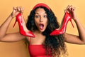 Young latin woman holding red high heel shoes afraid and shocked with surprise and amazed expression, fear and excited face