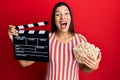 Young latin woman holding video film clapboard and popcorn celebrating crazy and amazed for success with open eyes screaming Royalty Free Stock Photo