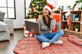 Young latin woman using laptop sitting by christmas tree smiling with hand over ear listening an hearing to rumor or gossip