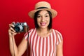 Young latin woman wearing summer hat holding vintage camera looking positive and happy standing and smiling with a confident smile Royalty Free Stock Photo