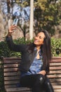 Young latina woman taking selfie on cell phone, resting on park bench on spring day.