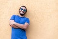 Young latino man with glasses, crossed arms and long hair, smiling looking at the camera, leaning against a yellow wall Royalty Free Stock Photo