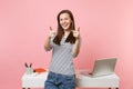 Young laughing woman pointing index fingers on camera work standing near white desk with laptop isolated on pastel pink Royalty Free Stock Photo