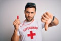 Young lifeguard man with beard wearing t-shirt with red cross and sunglasses using whistle with angry face, negative sign showing
