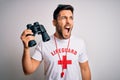 Young lifeguard man with beard wearing t-shirt with red cross and sunglasses using whistle angry and mad screaming frustrated and