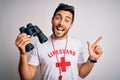 Young lifeguard man with beard wearing t-shirt with red cross and sunglasses using whistle cheerful with a smile on face pointing