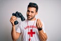 Young lifeguard man with beard wearing t-shirt with red cross and sunglasses using whistle doing money gesture with hands, asking