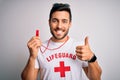 Young lifeguard man with beard wearing t-shirt with red cross and sunglasses using whistle happy with big smile doing ok sign,
