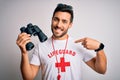 Young lifeguard man with beard wearing t-shirt with red cross and sunglasses using whistle looking confident with smile on face,