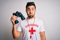 Young lifeguard man with beard wearing t-shirt with red cross and sunglasses using whistle puffing cheeks with funny face