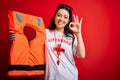 Young lifeguard woman holding rescue lifejacket over red background doing ok sign with fingers, excellent symbol
