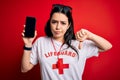 Young lifeguard woman showing smartphone screen over red background with angry face, negative sign showing dislike with thumbs