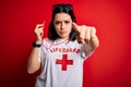 Young lifeguard woman wearing guard equipement holding whistle over red background pointing with finger to the camera and to you,