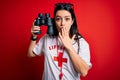 Young lifeguard woman wearing secury guard equipent holding binoculars over red background cover mouth with hand shocked with