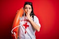 Young lifeguard woman wearing secury guard equipent holding rescue float over red background cover mouth with hand shocked with