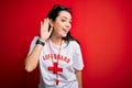 Young lifeguard woman wearing secury guard equipent over red background smiling with hand over ear listening an hearing to rumor