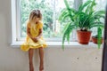 Young little blonde girl barefoot sitting on window sill thinking looking down near plant fern in clay pot. Side view. Royalty Free Stock Photo