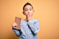 Young little boy kid eating sweet chocolate bar for dessert over isolated yellow background cover mouth with hand shocked with Royalty Free Stock Photo