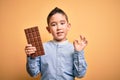 Young little boy kid eating sweet chocolate bar for dessert over isolated yellow background doing ok sign with fingers, excellent Royalty Free Stock Photo