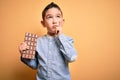 Young little boy kid eating sweet chocolate bar for dessert over isolated yellow background serious face thinking about question, Royalty Free Stock Photo