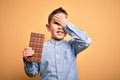 Young little boy kid eating sweet chocolate bar for dessert over isolated yellow background stressed with hand on head, shocked Royalty Free Stock Photo