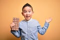 Young little boy kid eating sweet chocolate bar for dessert over isolated yellow background very happy and excited, winner Royalty Free Stock Photo