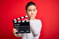 Young little boy kid filming video holding cinema director clapboard over isolated red background cover mouth with hand shocked