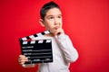Young little boy kid filming video holding cinema director clapboard over isolated red background serious face thinking about