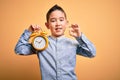 Young little boy kid holding classic bell alarm clock over isolated yellow background doing ok sign with fingers, excellent symbol Royalty Free Stock Photo