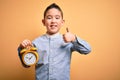 Young little boy kid holding classic bell alarm clock over isolated yellow background happy with big smile doing ok sign, thumb up Royalty Free Stock Photo