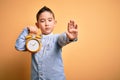 Young little boy kid holding classic bell alarm clock over isolated yellow background with open hand doing stop sign with serious Royalty Free Stock Photo