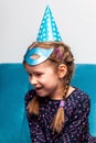 Young little bright cheerful happy birthday girl in a blue mask and a party hat smiling, solo portrait. Ponytails, smile