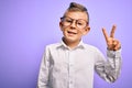 Young little caucasian kid with blue eyes wearing glasses and white shirt over purple background smiling with happy face winking Royalty Free Stock Photo