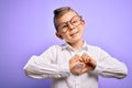 Young little caucasian kid with blue eyes wearing glasses and white shirt over purple background smiling in love showing heart Royalty Free Stock Photo