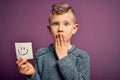 Young little caucasian kid showing smiley face on a paper note as happy message cover mouth with hand shocked with shame for
