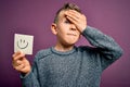 Young little caucasian kid showing smiley face on a paper note as happy message stressed with hand on head, shocked with shame and
