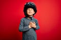 Young little caucasian kid wearing vintage biker motorcycle helmet and googles over red background smiling with hands on chest