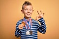 Young little caucasian winner kid wearing award competition medals over yellow background doing ok sign with fingers, excellent