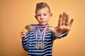 Young little caucasian winner kid wearing award competition medals over yellow background with open hand doing stop sign with