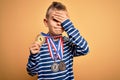 Young little caucasian winner kid wearing award competition medals over yellow background stressed with hand on head, shocked with